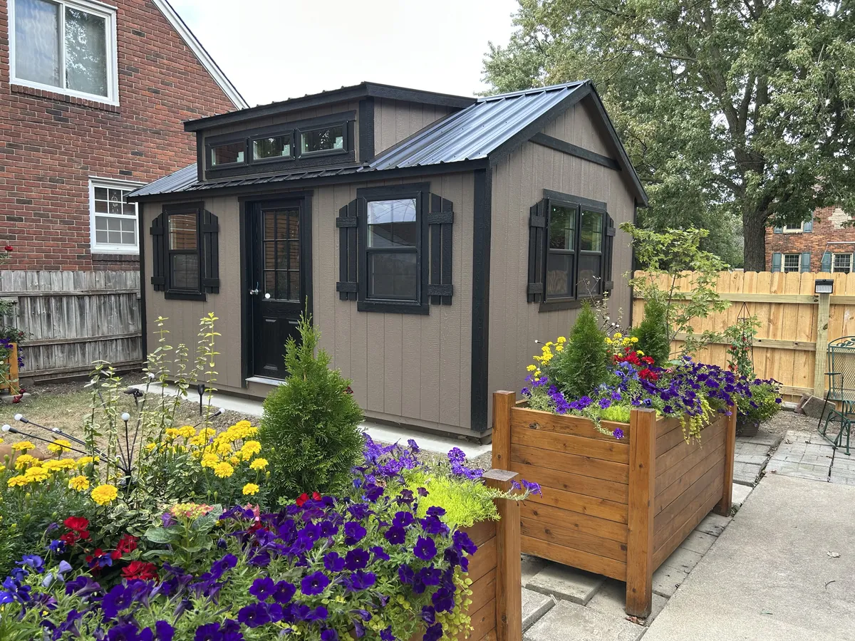 Brown studio shed with black trim, shutters, and transom windows behind a flower garden and cedar planter.