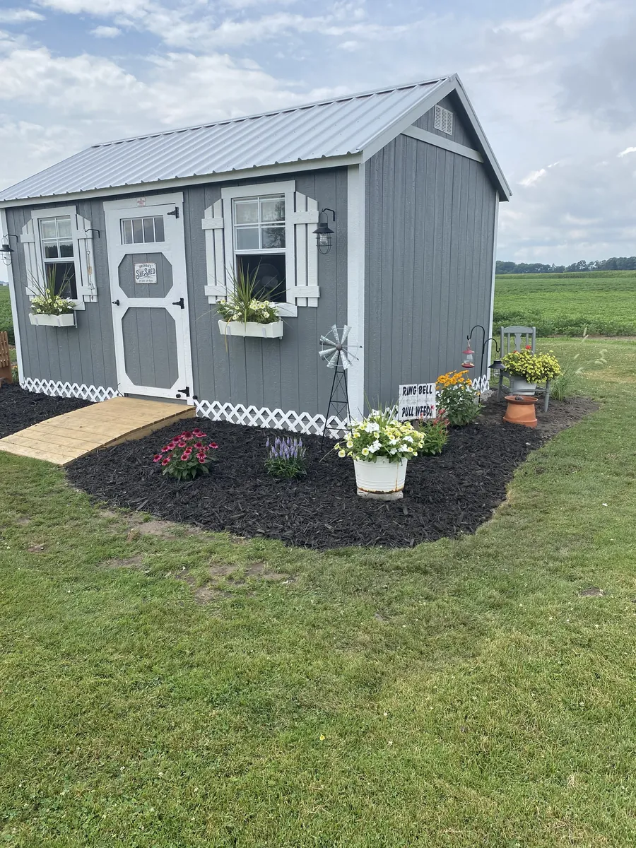 Gray garden shed with white trim, window flower boxes, and landscaped flower beds beside farm fields.